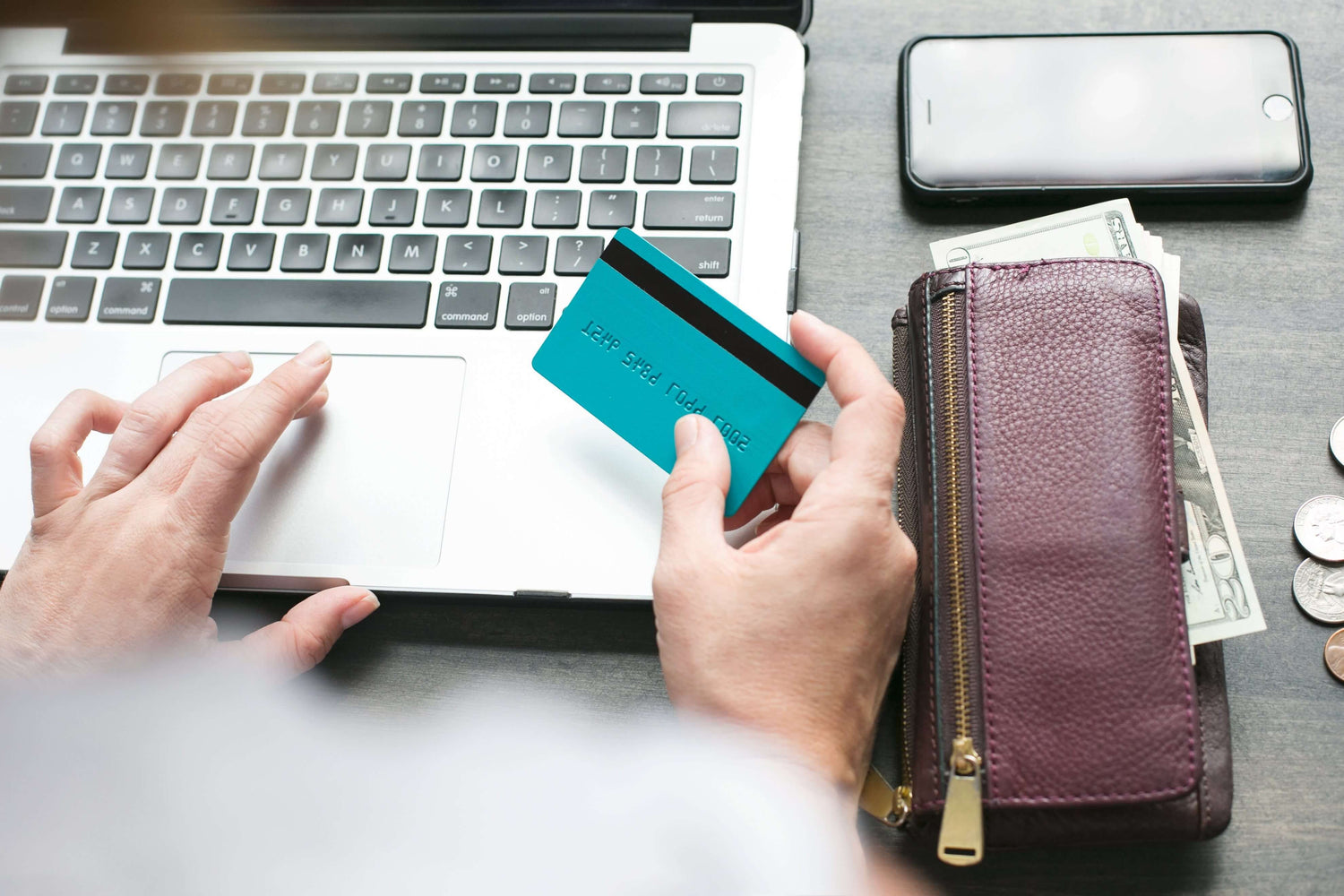 Person holding a teal card near a laptop with a wallet and phone on a table
