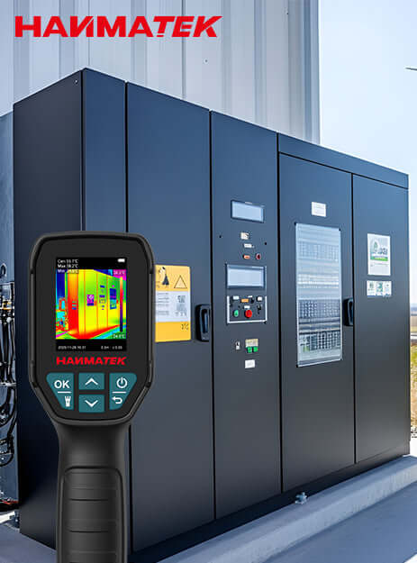 Handheld thermal imaging camera displaying temperature scan of industrial electrical control cabinets outdoors.