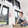 Person using a laser distance meter to measure height in a well-lit indoor space with large windows