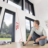Person using a laser distance meter to measure height in a well-lit indoor space with large windows