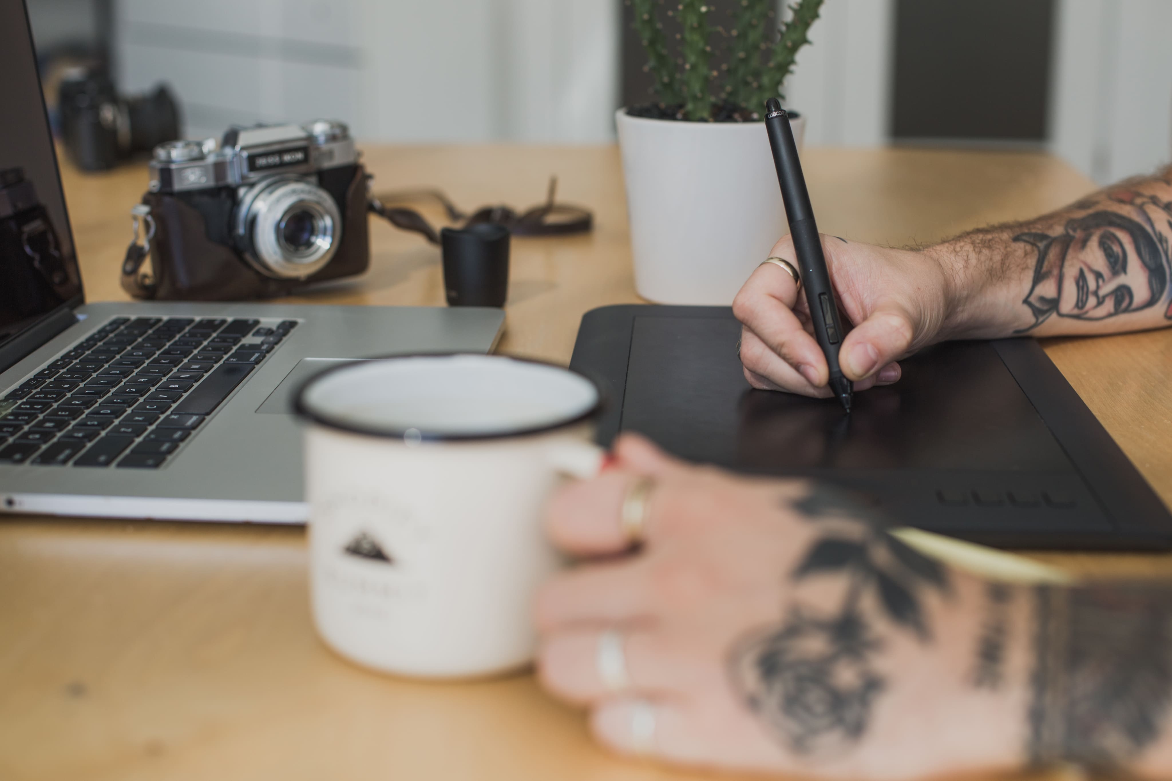 Person using a graphic tablet with tattoos on a desk next to a laptop and mug.