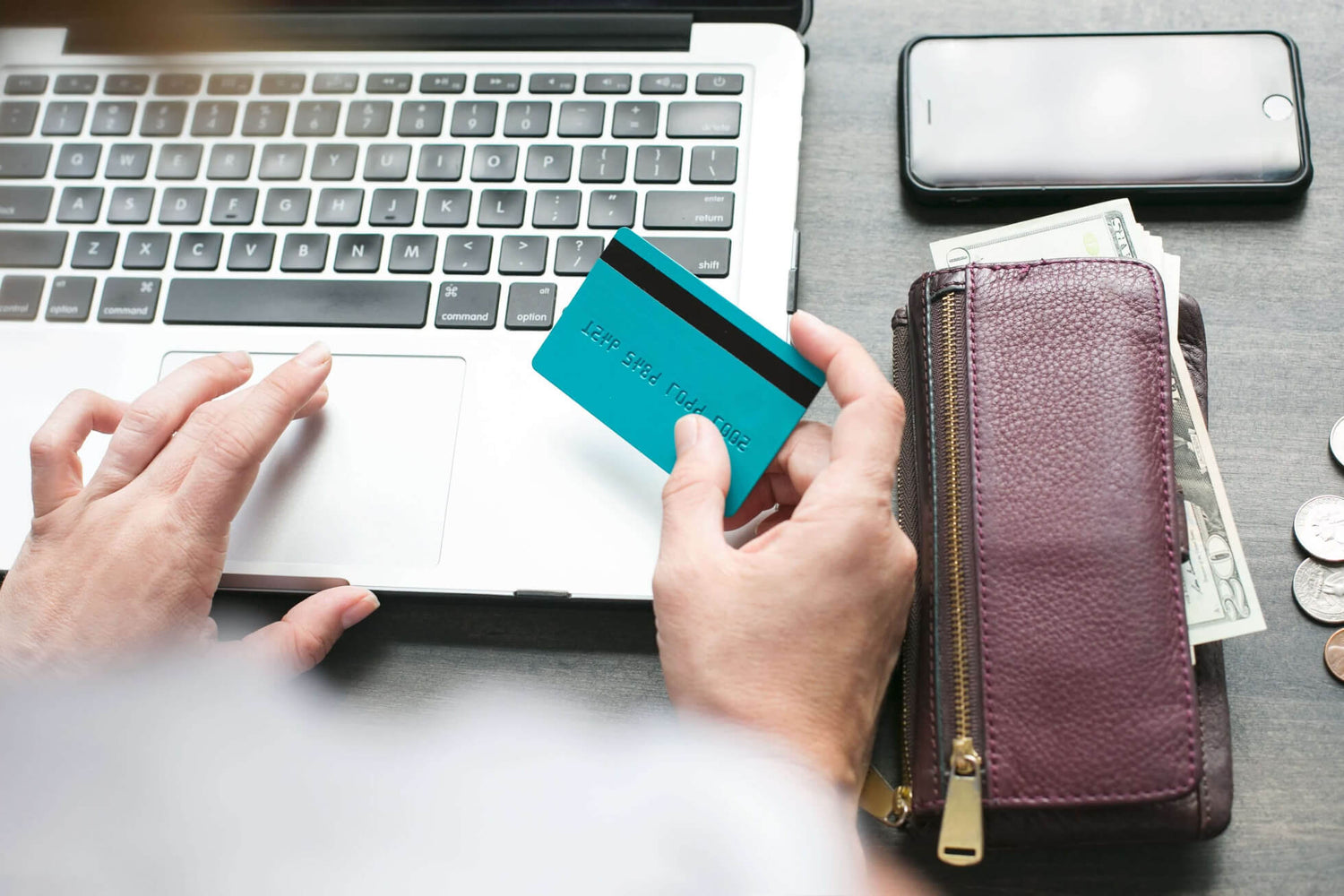 Person holding a teal card near a laptop with a wallet and phone on a table