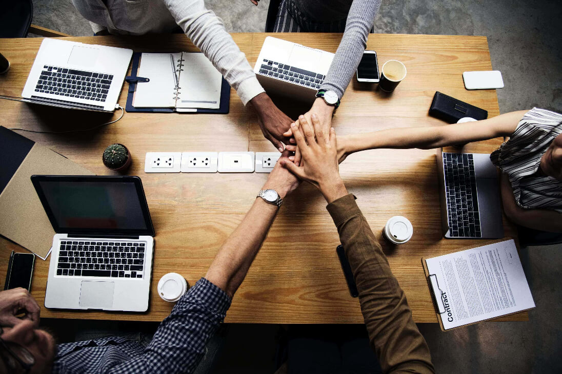 Group of people with hands stacked together at a wooden desk with laptops and office supplies.